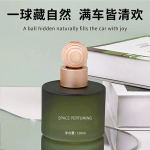 A dark green cylindrical car fragrance bottle with a round wooden-patterned cap and metallic gold collar, labeled 'SPACE PERFUMING' and '120ml'. The bottle is styled on a glass surface with books in the background, under Chinese and English text that reads 'A ball hidden naturally fills the car with joy'.