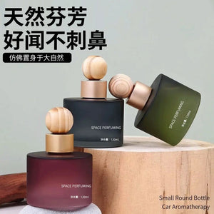 Three small round car aroma bottles with wooden caps displayed on a wooden pedestal against a pale green background. The bottles are in different colors: dark green, dark blue, and deep purple, with one bottle lying on its side.