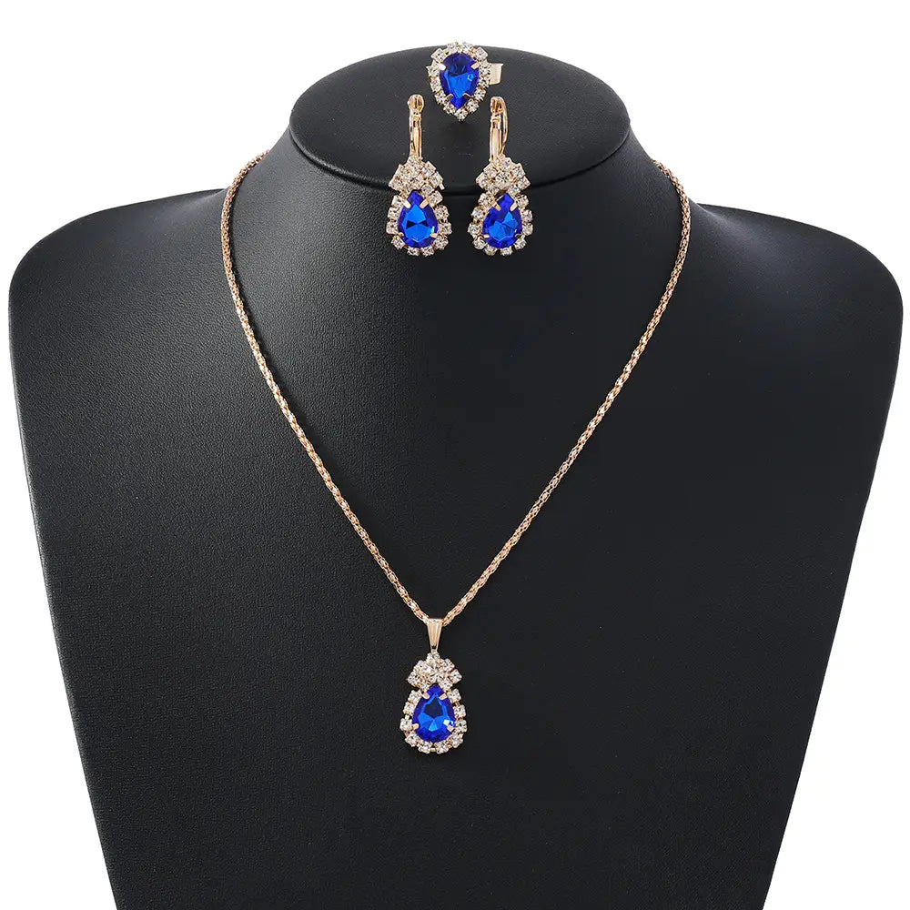 A gold-tone jewelry set featuring a teardrop-shaped pendant necklace and matching drop earrings, both adorned with vivid blue gemstones and clear rhinestone halos, displayed on a black jewelry bust against a white background.