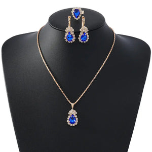 A gold-tone jewelry set featuring a teardrop-shaped pendant necklace and matching drop earrings, both adorned with vivid blue gemstones and clear rhinestone halos, displayed on a black jewelry bust against a white background.