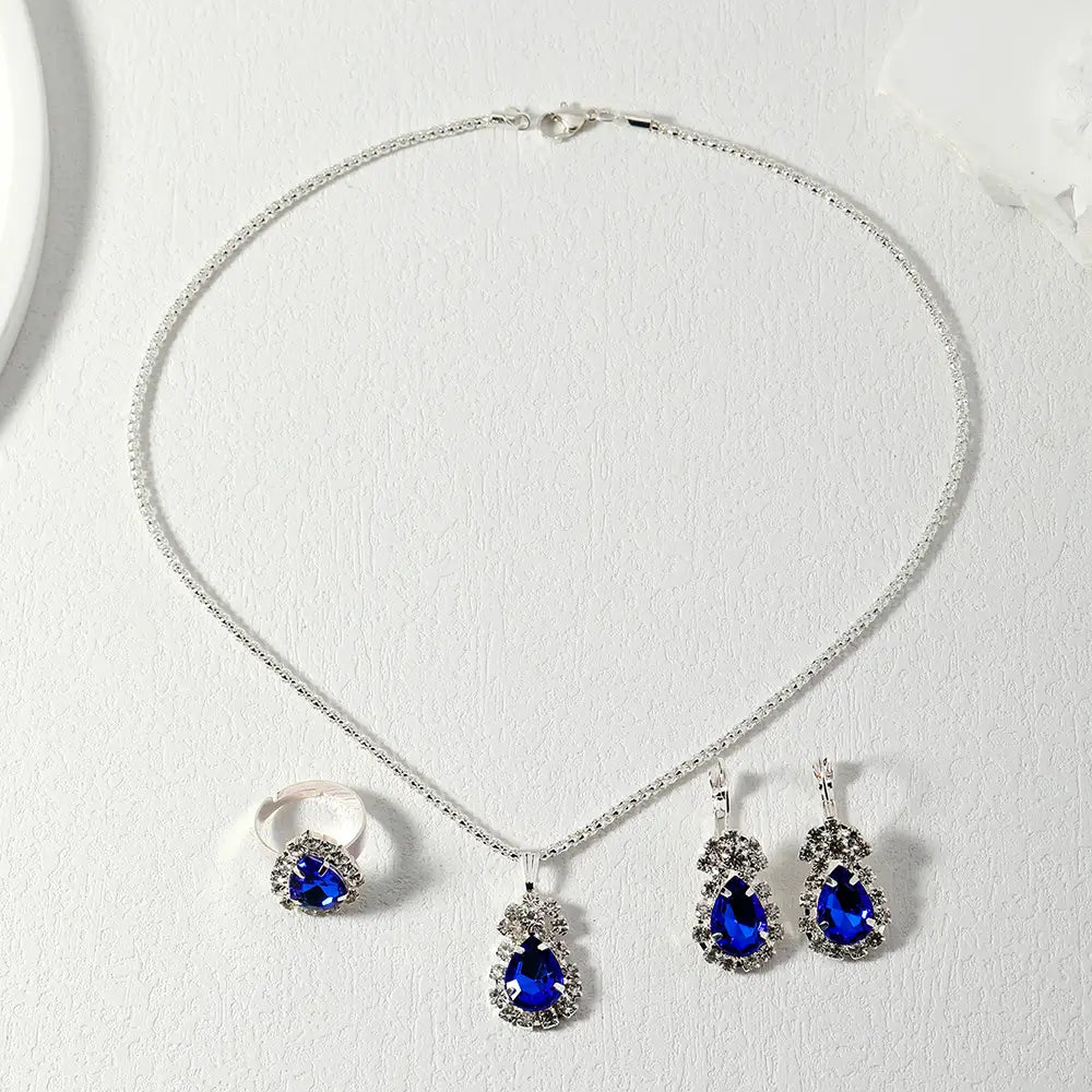 Silver-tone jewelry set featuring a necklace, ring, and earrings with deep blue teardrop-shaped crystals surrounded by clear rhinestones, displayed on a textured white surface.