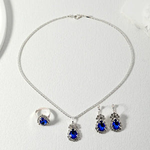 Silver-tone jewelry set featuring a necklace, ring, and earrings with deep blue teardrop-shaped crystals surrounded by clear rhinestones, displayed on a textured white surface.