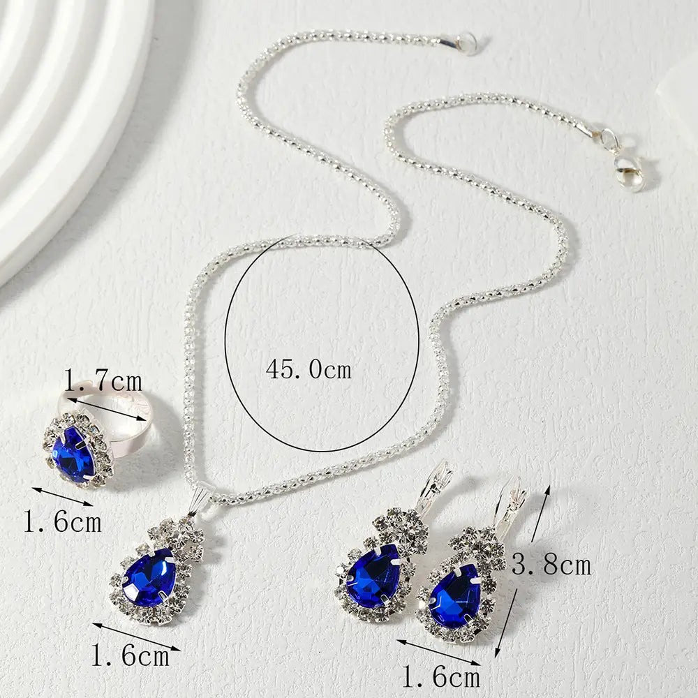 Silver-tone jewelry set with a necklace, ring, and earrings featuring deep blue teardrop-shaped crystals, shown with measurement labels on a light textured background.