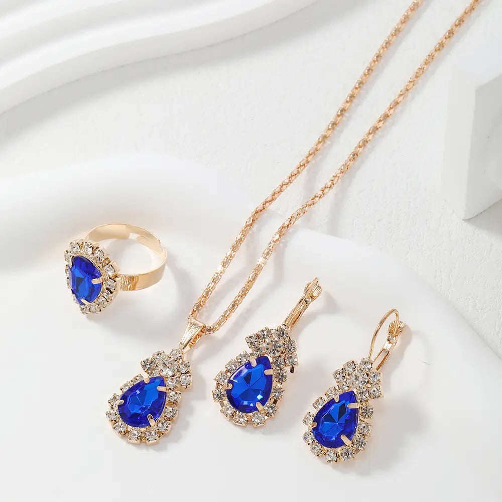 A gold jewelry set featuring a ring, necklace, and drop earrings with teardrop-shaped blue gemstones surrounded by clear crystals, displayed on a white background.