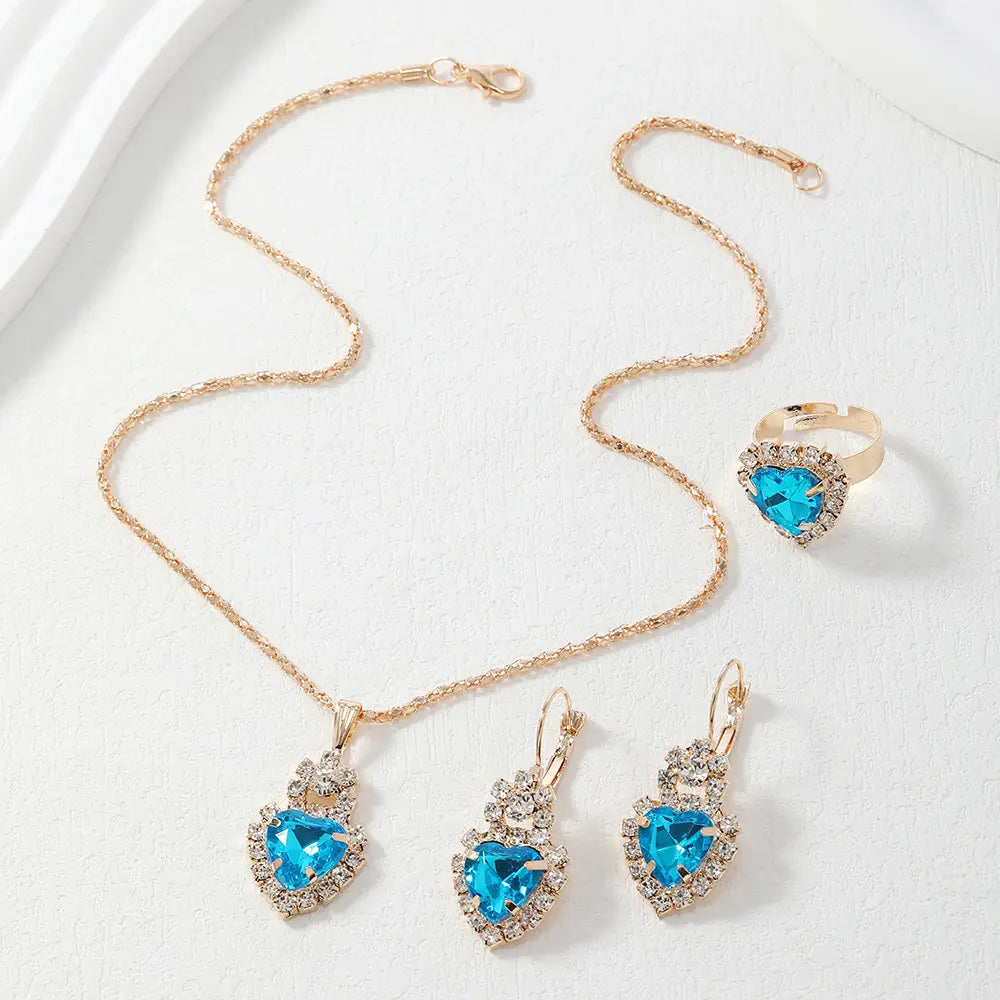 A gold-tone jewelry set featuring a heart-shaped pendant necklace, matching drop earrings, and a ring, all adorned with bright blue heart-shaped crystals surrounded by clear rhinestones, displayed on a textured white surface.