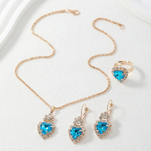 A gold-tone jewelry set featuring a heart-shaped pendant necklace, matching drop earrings, and a ring, all adorned with bright blue heart-shaped crystals surrounded by clear rhinestones, displayed on a textured white surface.