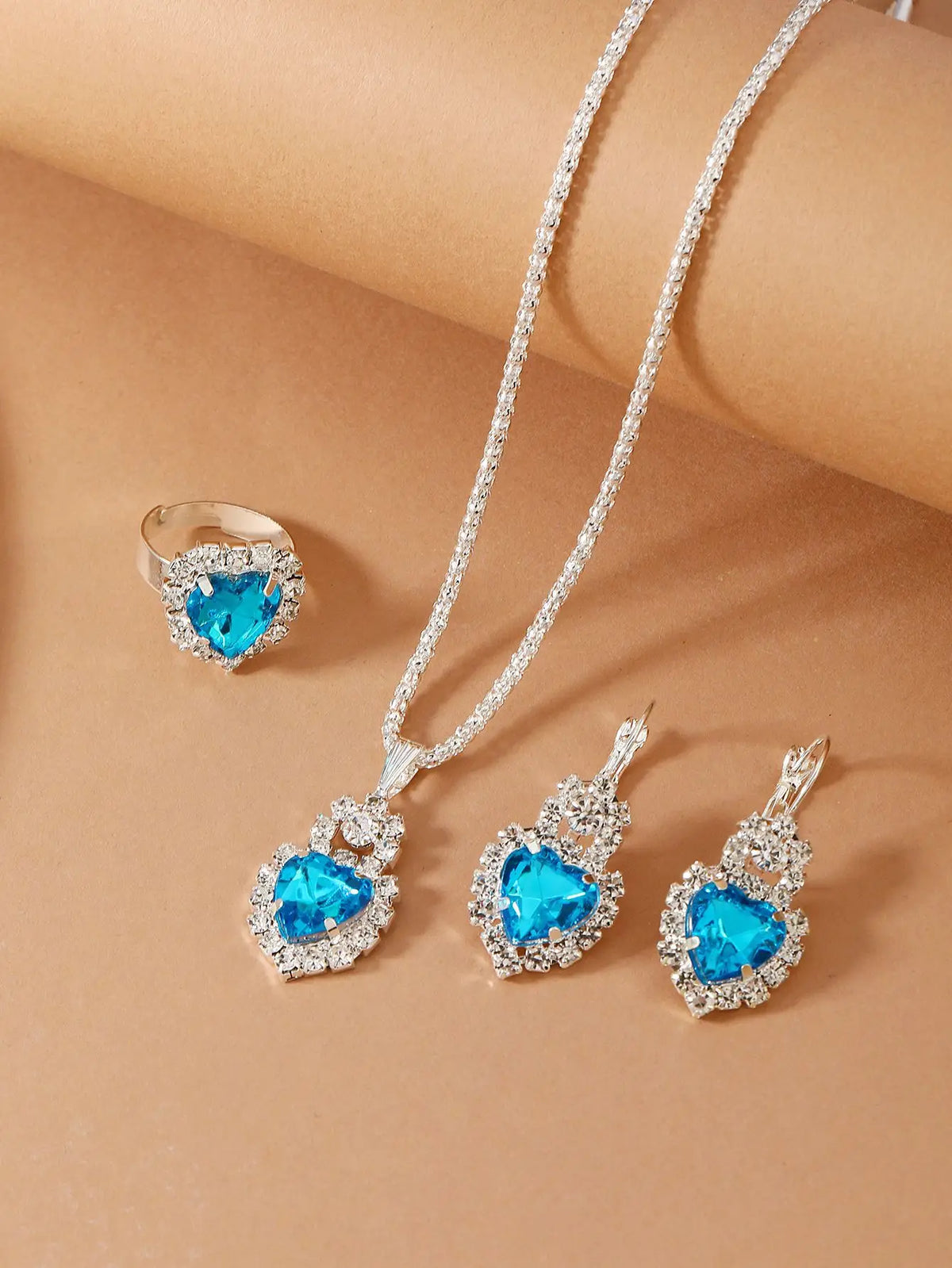 Silver-tone jewelry set featuring a heart-shaped blue gemstone pendant necklace, matching drop earrings, and a ring, displayed on a warm beige background.